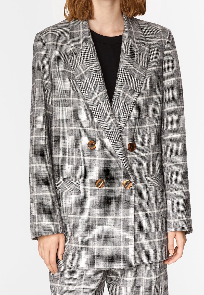 Femme portant un blazer croisé à carreaux gris avec des boutons marron sur un top noir et un pantalon à carreaux assorti.
