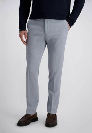 Man draagt lichtgrijze pantalon, donkerbruine leren loafers en een zwart overhemd met lange mouwen, met handen in de zakken.