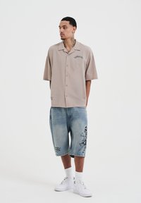 Lichtbeige kortekshorts overhemd met knoopjes en kraag; lichtblauwe knielange denim shorts met zwart grafisch patroon; witte sneakers.