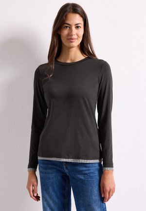 Femme aux longs cheveux foncés portant une chemise noire à manches longues avec une bordure à carreaux et un jean bleu, debout devant un fond blanc.