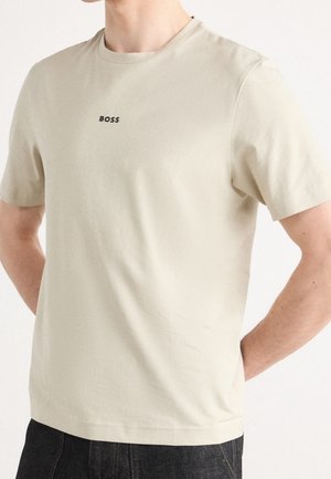 Mann in beiger Kurzarm-T-Shirt mit schwarzem "BOSS"-Logo, das mittig auf der Brust platziert ist, kombiniert mit dunklen Jeans.