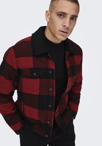 Jachetă cu carouri roșii și negre, având un guler din fleece negru, nasturi cu prindere prin apăsare și buzunare pe piept. Fabricată din bumbac, cu un design clasic pentru activități în aer liber.