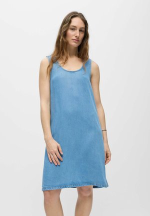 Femme aux longs cheveux portant une robe en jean bleu clair sans manches jusqu'aux genoux, sur un fond clair uni.