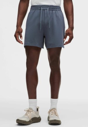 Mand iført blå træningsshorts, hvide mellemlange sokker og beige løbesko, står mod en ensfarvet baggrund.