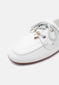Mocasín de cuero blanco con cordones grises atados en un lazo, con charms dorados en forma de corazón y cuentas junto a los ojales y borde de la suela marrón.