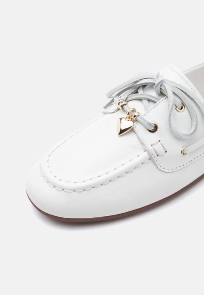 Mocassin en cuir blanc avec lacets gris noués en nœud, ornés de charms en forme de cœur doré et de perles près des œillets, avec bordure de semelle marron.