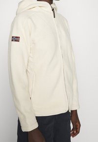 Persona che indossa un felpa con zip color crema e cappuccio, con una toppa della bandiera della Norvegia sulla manica superiore, abbinata a pantaloni scuri.