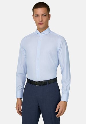 Uomo che indossa una camicia azzurra a maniche lunghe, pantaloni blu scuro e cintura nera, in piedi davanti a uno sfondo uniforme.