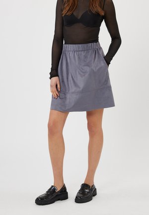 Femme portant un haut en dentelle noir à manches longues, une jupe grise au genou avec poches, et des mocassins noirs épais, se tenant sur un fond blanc.