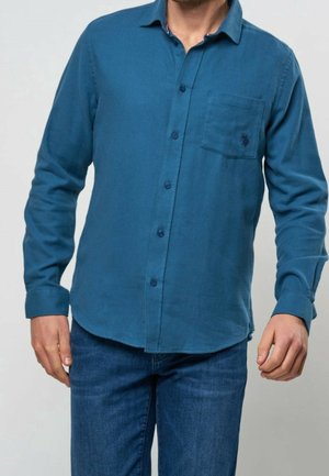 Mann trägt ein langärmeliges türkisfarbenes Hemd mit Brusttasche und blaue Jeans, steht vor einem einfarbigen hellen Hintergrund.