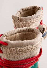Coppia di stivali invernali beige foderati in pile con lacci rossi e linguette di tessuto su sfondo neutro.