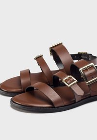 Sandalias de cuero marrón que cuentan con múltiples correas ajustables, hebillas en tono dorado y una suela plana con un borde texturizado para mayor agarre.