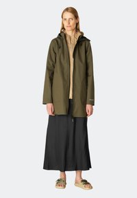Olijfgroene waterdichte jas met een ritssluiting aan de voorkant, een verstelbare capuchon en twee zijzakken. Draagt over een beige top en een zwarte rok.
