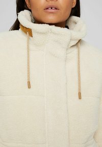 Kermanvärinen fleece-takki, jossa on korkea kaulus, ja kaulassa on viininpunainen corduroy-yllätys sekä kultasävyiset nyörit. Pehmeä tekstuuri, palotettu muotoilu.