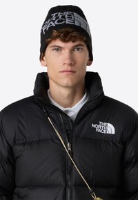 Giacca a vento nera con colletto alto, chiusura a zip e logo. Berretto in maglia nero con logo bianco. L'uomo indossa una polo bianca sotto.