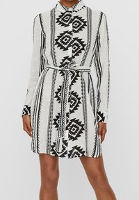 Långärmad skjortklänning i lätt, strukturerad tyg. Har ett djärvt svart geometriskt mönster på en vit bakgrund. Justerbart bälte.