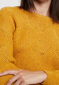 Pletený žlutý svetr s texturovaným vzorem, který obsahuje geometrické tvary, žebrovaný výstřih a manžety a barevné tečky po celém povrchu.