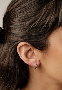 Srebrny kolczyk w kształcie obręczy na uchu modelki, z gładką, wypolerowaną powierzchnią i zaokrąglonym, kroplowym kształtem. Częściowo widoczne włosy.