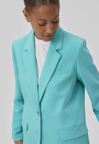 Turquoise blazer met notch revers, sluiting met één knoop, voorzakken en een gladde stofstructuur. Gedragen over een wit shirt.