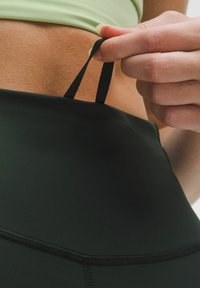 Czarne legginsy o wysokim stanie z gładkiego, elastycznego materiału. Widoczne szwy oraz ręka ciągnąca za pętelkę sznurka w pasie.