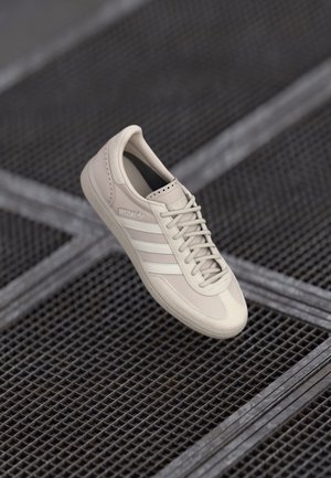 Zapatilla Adidas Spezial beige con rayas y cordones blancos flotando sobre una superficie de rejilla metálica.