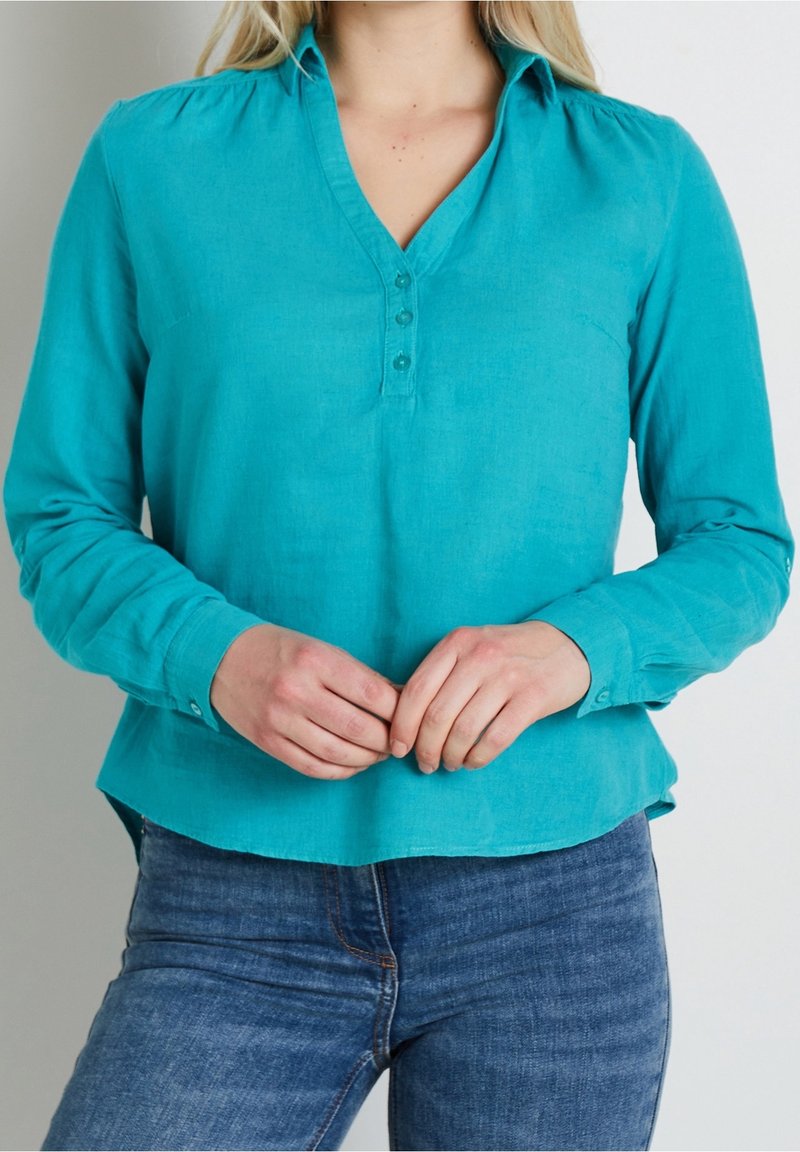 Femme portant un chemisier bleu sarcelle à manches longues avec trois boutons et un jean bleu, les mains jointes devant, sur un fond uni.
