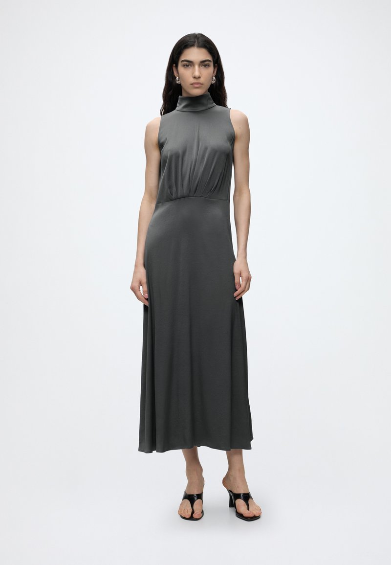 Femme aux longs cheveux foncés portant une robe midi gris foncé sans manches à col montant et des sandales à talons ouvertes noires, debout devant un fond uni.