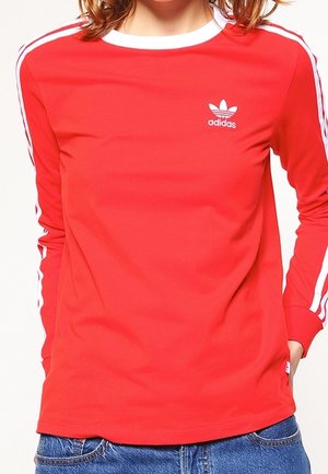 Femme portant un haut rouge à manches longues Adidas avec des bandes blanches sur les manches et le logo Adidas sur la poitrine.