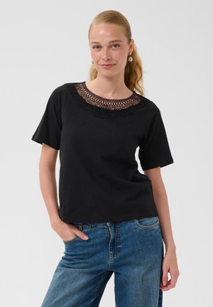 Kobieta z blond kucykiem, ubrana w czarną koszulkę z krótkim rękawem z koronkowym dekoltem i niebieskie dżinsy, stoi z jedną ręką w kieszeni.