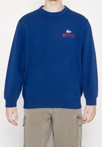 Modrá mikina s žebrovaným kulatým výstřihem, s červeným nápisem "NETFLIX" a šedým logem krokodýla. Dlouhé rukávy a volný střih.