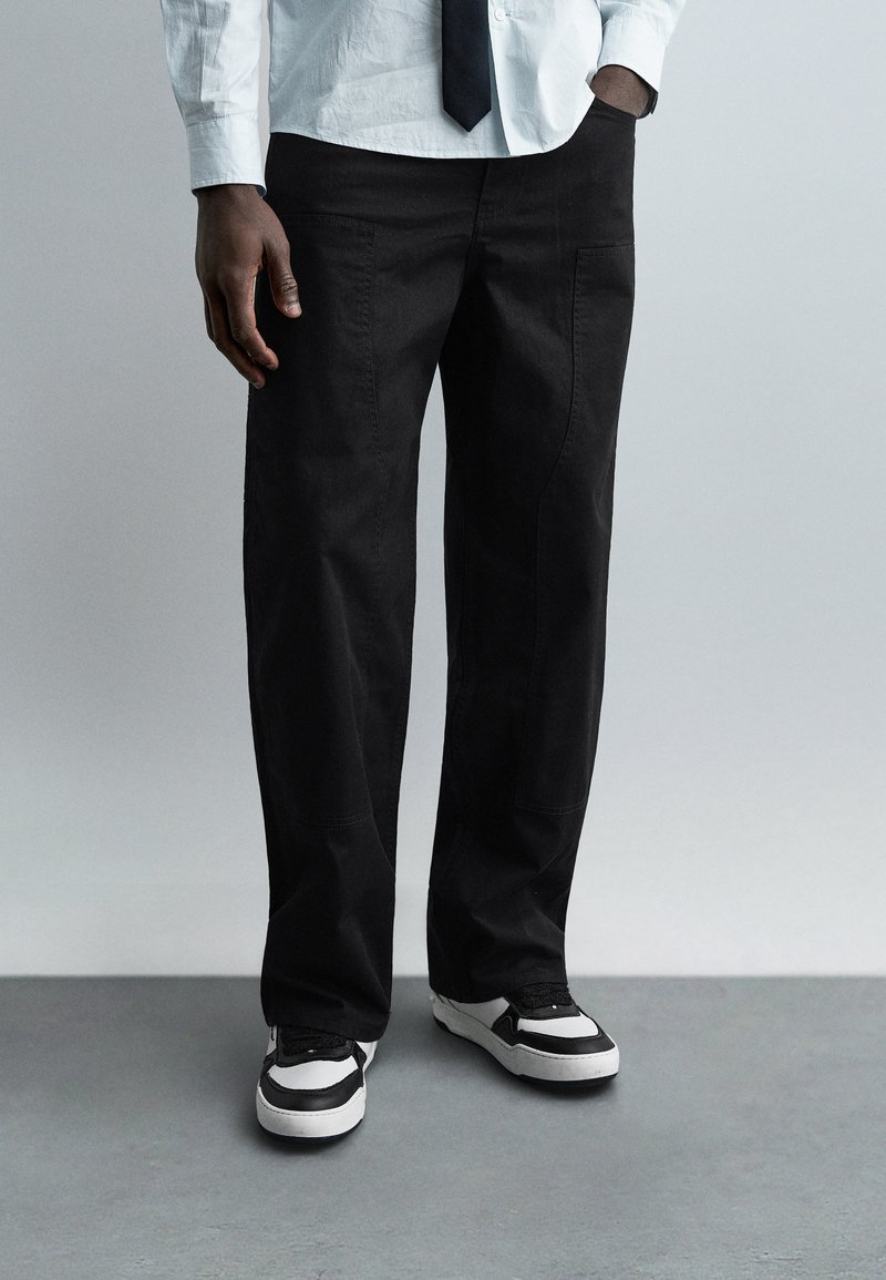 Άνδρας με μαύρο παντελόνι, μαύρα και λευκά αθλητ ικά παπούτσια, λευκό πουκάμισο και μαύρη γραβάτα, στέκεται με το ένα χέρι στην τσέπη σε γκρι πάτωμα.