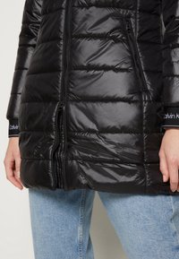Ingrandimento di una persona che indossa una giacca trapuntata nera con zip frontale e tasche laterali, abbinata a un paio di jeans in denim blu chiaro.