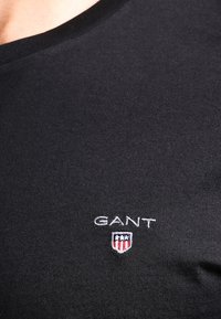 Black cotton t-shirt with a round neckline featuring a white embroidered "GANT" logo and a small shield design in red, white, and blue.