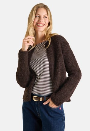 Blonde woman smiling wearing a dark brown fuzzy cardigan over a black and white striped shirt with blue jeans and a black belt with a gold buckle.