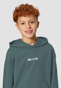 Tirkīza kapuce ar kangaroo kabatu un saites. Uz krūtīm atrodas balts "SLVR" logotips. Mīksts, teksturēts audums ar brīvu piegriezumu.