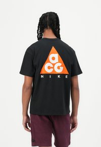 Schwarzes Baumwoll-T-Shirt mit einem großen orangefarbenen dreieckigen Logo und weißem Text "acg" und "NIKE" auf der Rückseite. Kurze Ärmel und entspannte Passform.