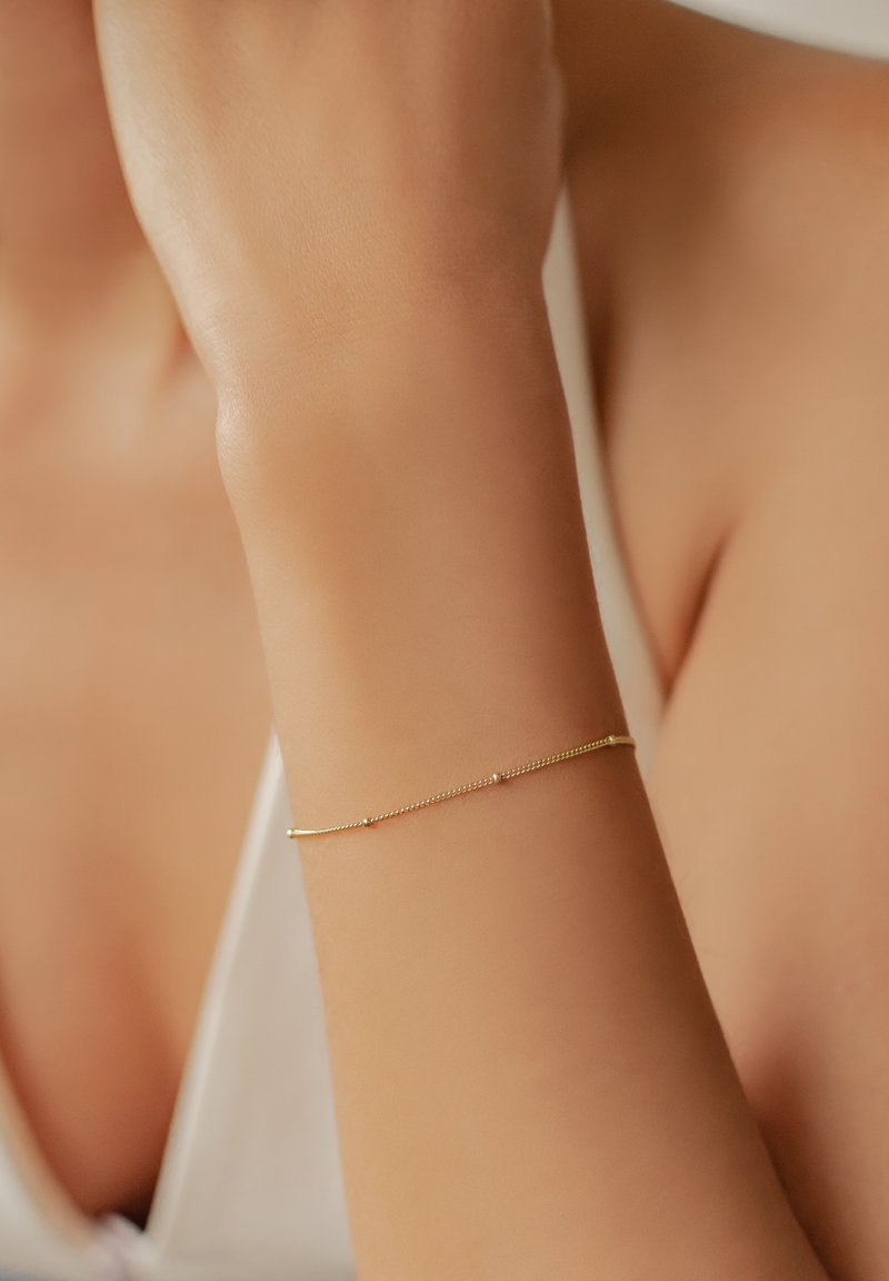 Pulsera de oro con un diseño de cadena delicada, que presenta pequeños acentos coloridos y una textura suave, llevada en una muñeca.