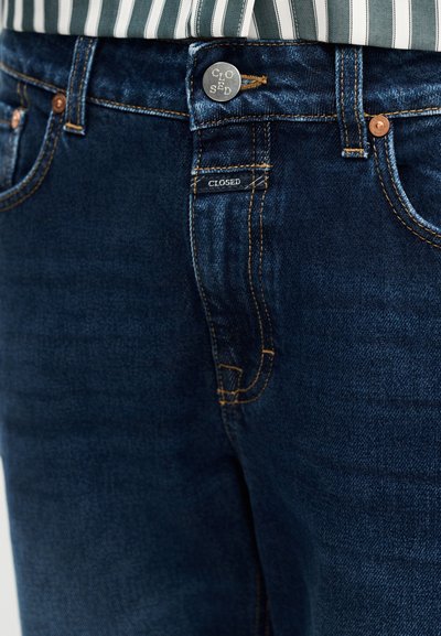 Σκούρο μπλε τζιν με ελαφρώς κωνική εφαρμογή, με κλείσιμο με κουμπιά, πορτοκαλί ραφές και εμφανές λογότυπο στο μπροστινό μέρος.