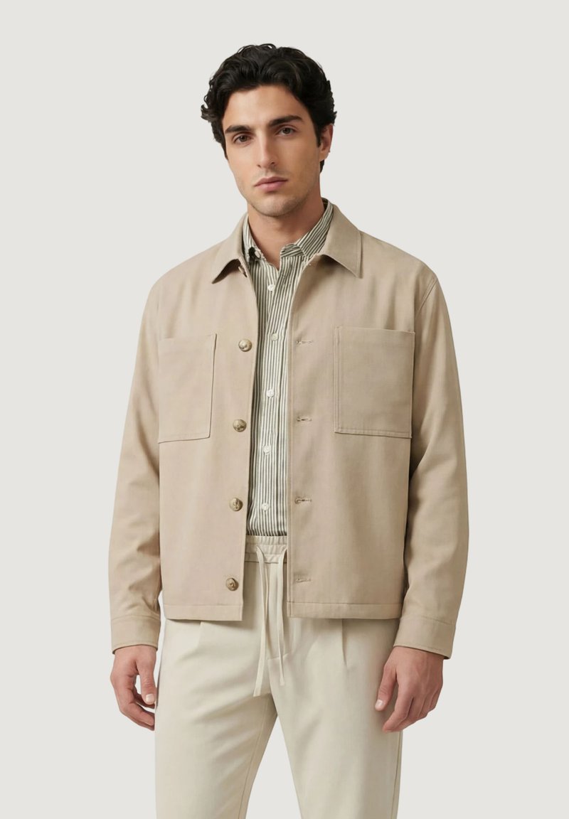 Homme portant une veste beige boutonnée sur une chemise à rayures claires et un pantalon crème, debout devant un fond uni.
