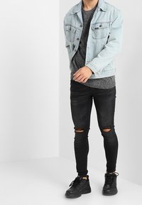 Chaqueta de mezclilla azul claro con dos bolsillos en el pecho, suéter texturizado gris oscuro debajo, jeans skinny negros rasgados y zapatillas negras.