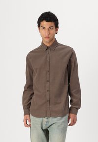 Braunes Hemd mit Knöpfen aus weichem Stoff, mit Kragen, langen Ärmeln und geradem Schnitt. Getragen mit hellblauen Jeans.