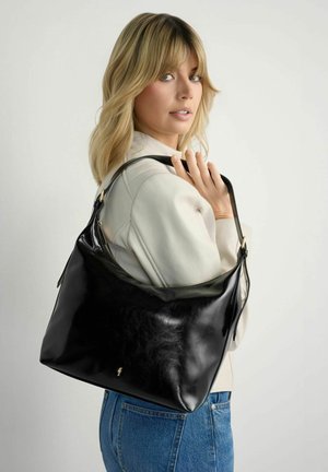 Blondynka w białej kurtce i niebieskich dżinsach trzyma dużą czarną skórzaną torbę na ramię na jednolitym tle.