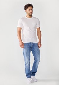Hvit teksturert t-skjorte med en liten lomme, paret med lyseblå jeans. Modellen har på seg hvite joggesko, stående foran en ensfarget bakgrunn.