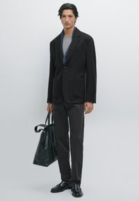 Blazer nero in lana con due tasche frontali, abbinato a una maglietta grigia a collo rotondo, pantaloni scuri e una borsa a tracolla nera in pelle. Scarpe nere eleganti.