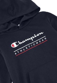 Felpa con cappuccio blu navy presenta una tasca frontale e il logo Champion bianco/rosso con il testo "ATHLETICWEAR" sul petto. Texture liscia.