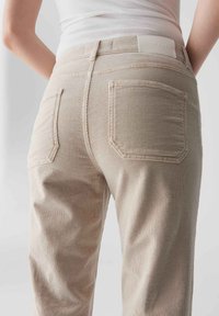 Beige Cord-Hosen mit zwei Gesäßtaschen, sichtbaren Nähdetails und einem Etikett am Bund. Der Stoff zeigt feine vertikale Streifen und Textur.