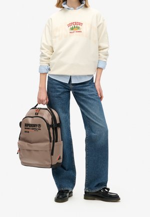 Beige sweatshirt met "SUPERDRY" logo, lichtblauw shirt eronder, denim flared jeans en een brown rugzak met zwarte accenten en logo.