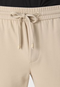 Beige sweatpants med en tekstureret overflade, elastisk talje og justerbar snørre med cremefarvede spidser. En enkelt sidelomme synlig.