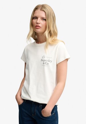 Ung kvinde med langt blondt hår iført en hvid Superdry T-shirt og mørkeblå jeans med hænderne i lommerne, mod en ensfarvet baggrund.
