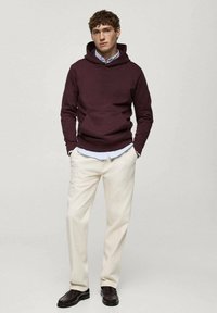 Burgundowa bluza z kapturem z przednią kieszenią, noszona na jasnoniebieskiej koszuli. Połączona z kremowymi spodniami i ciemnymi butami. Model stoi z rękami w kieszeniach.